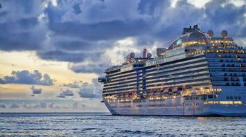 A large cruise ship sailing on calm waters during sunset, with illuminated decks and a cloudy sky in the background