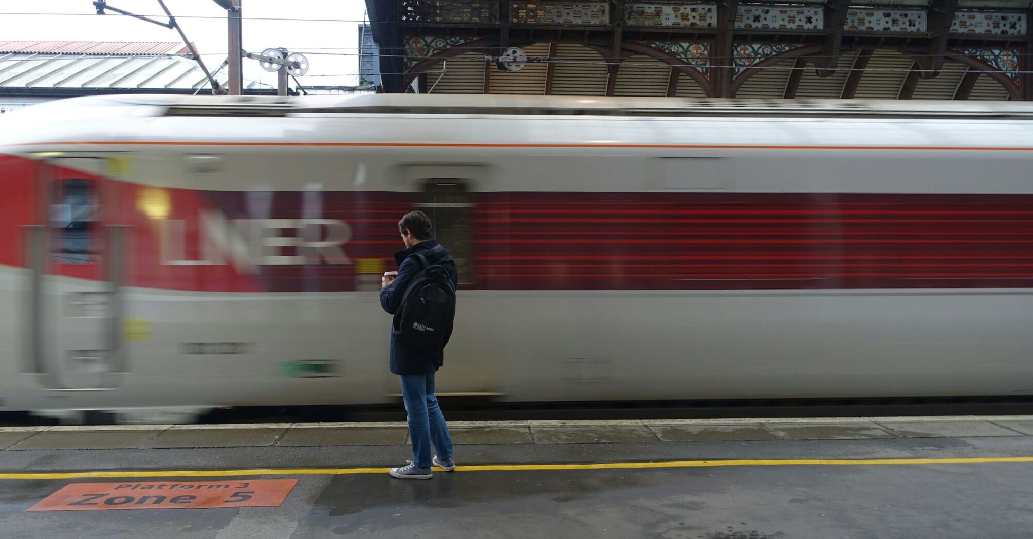 A man stands on a train platform as an LNER train speeds past, creating a blurred motion effect