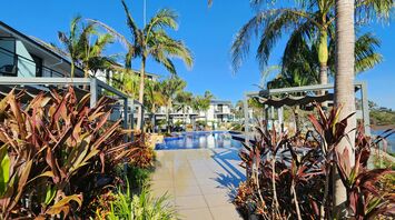 Outdoor pool area surrounded by tropical plants and modern resort buildings under a clear blue sky