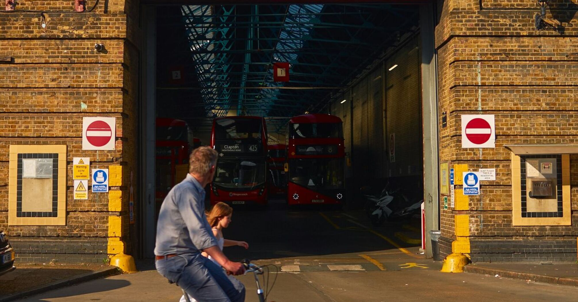 A cyclist with a child passes by a Stagecoach depot with several red buses parked inside