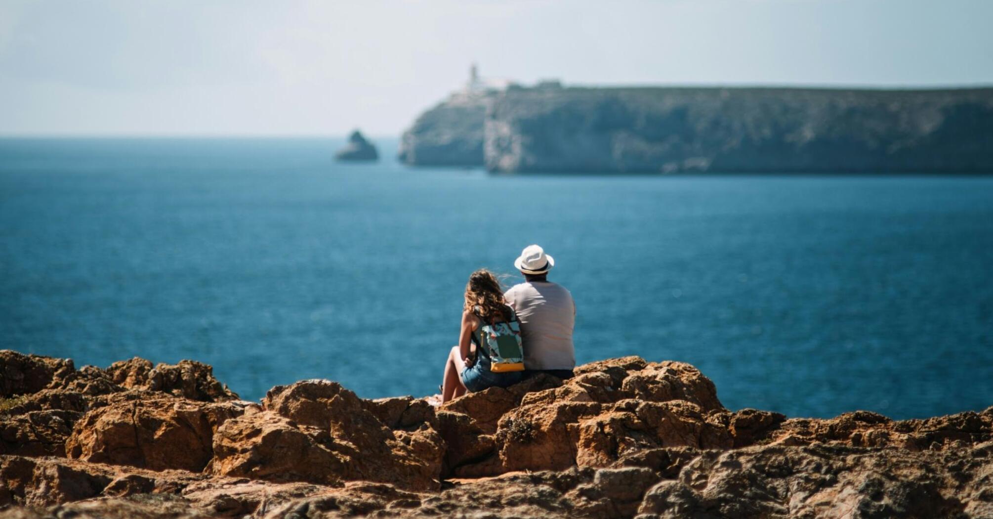 A couple sitting on rocky cliffs overlooking the ocean at Sagres, Portugal