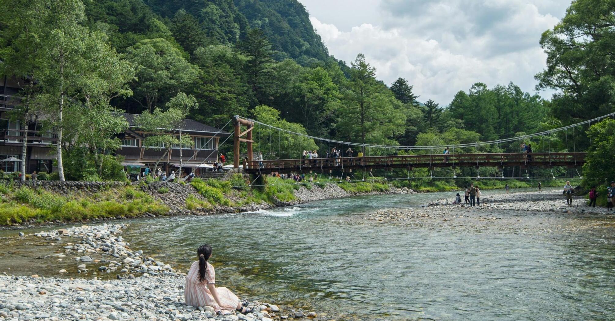 A person in a pink dress sitting by a river in Kamikochi, with a lush forested mountain backdrop and a suspension bridge with people walking on it