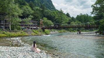 A person in a pink dress sitting by a river in Kamikochi, with a lush forested mountain backdrop and a suspension bridge with people walking on it