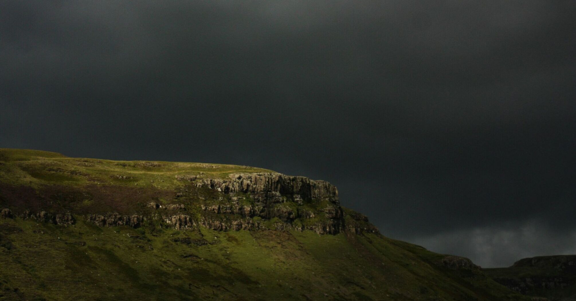 A dark and stormy sky looms over a rugged green hill with rocky outcrops, hinting at an impending storm