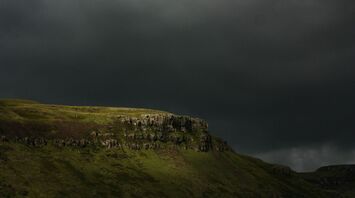 A dark and stormy sky looms over a rugged green hill with rocky outcrops, hinting at an impending storm