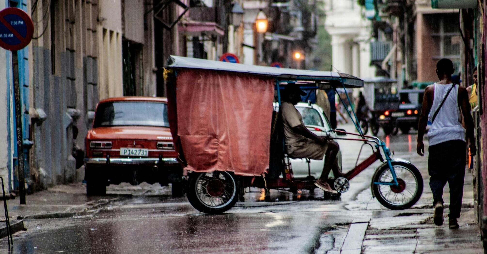 A rainy street in Havana with a tricycle taxi and an old car