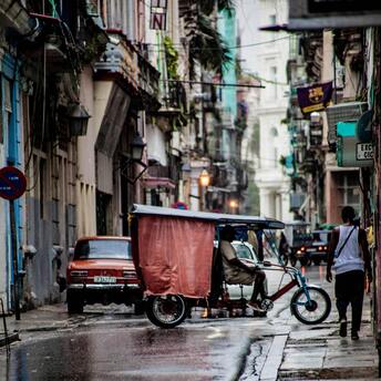 A rainy street in Havana with a tricycle taxi and an old car