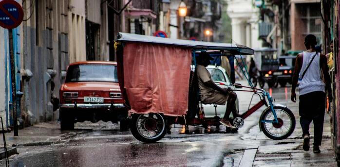A rainy street in Havana with a tricycle taxi and an old car