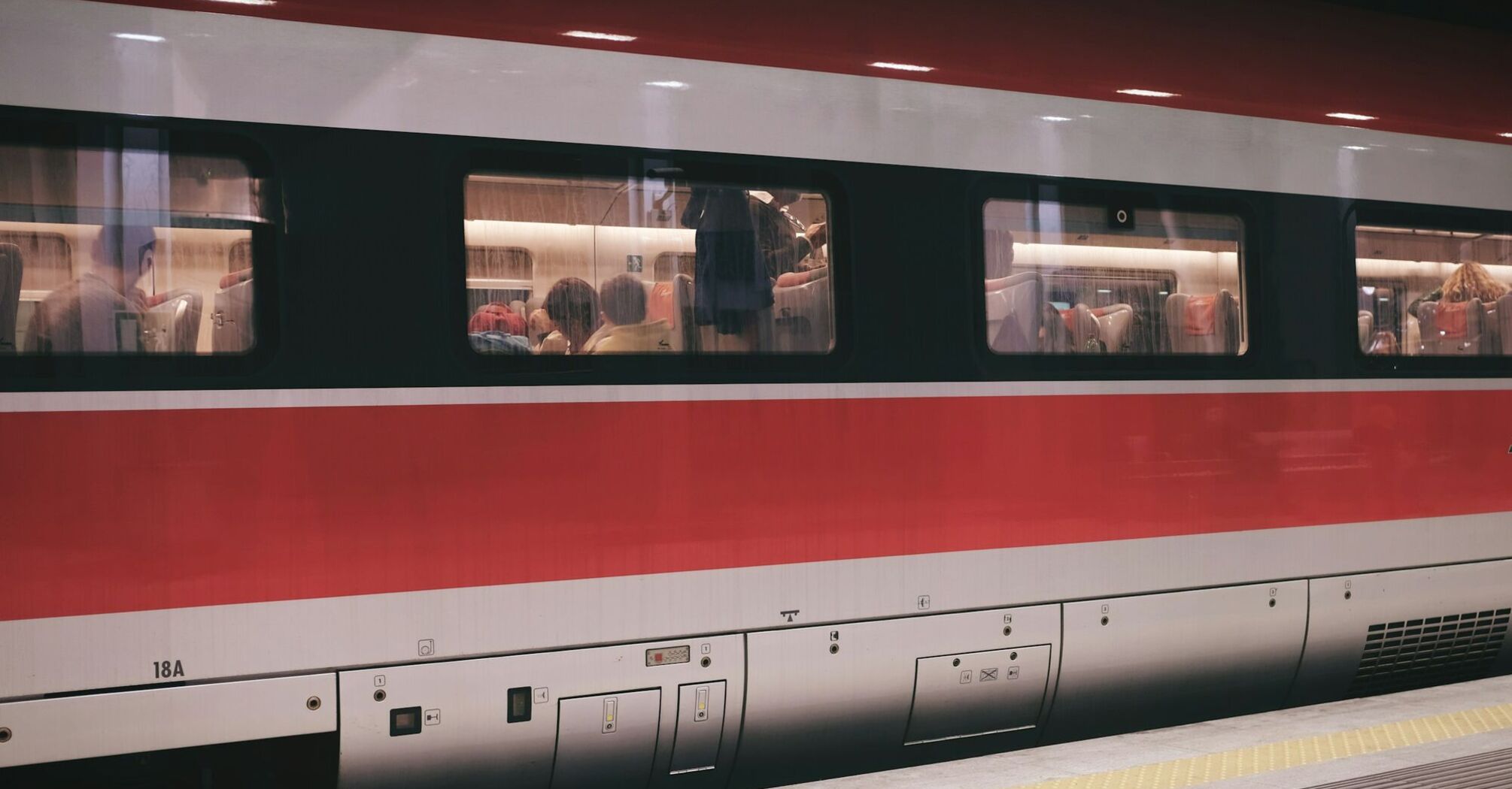 A red and white passenger train with people seated inside at a platform