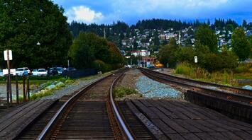 Railroad tracks extending towards a forested hillside town under cloudy skies