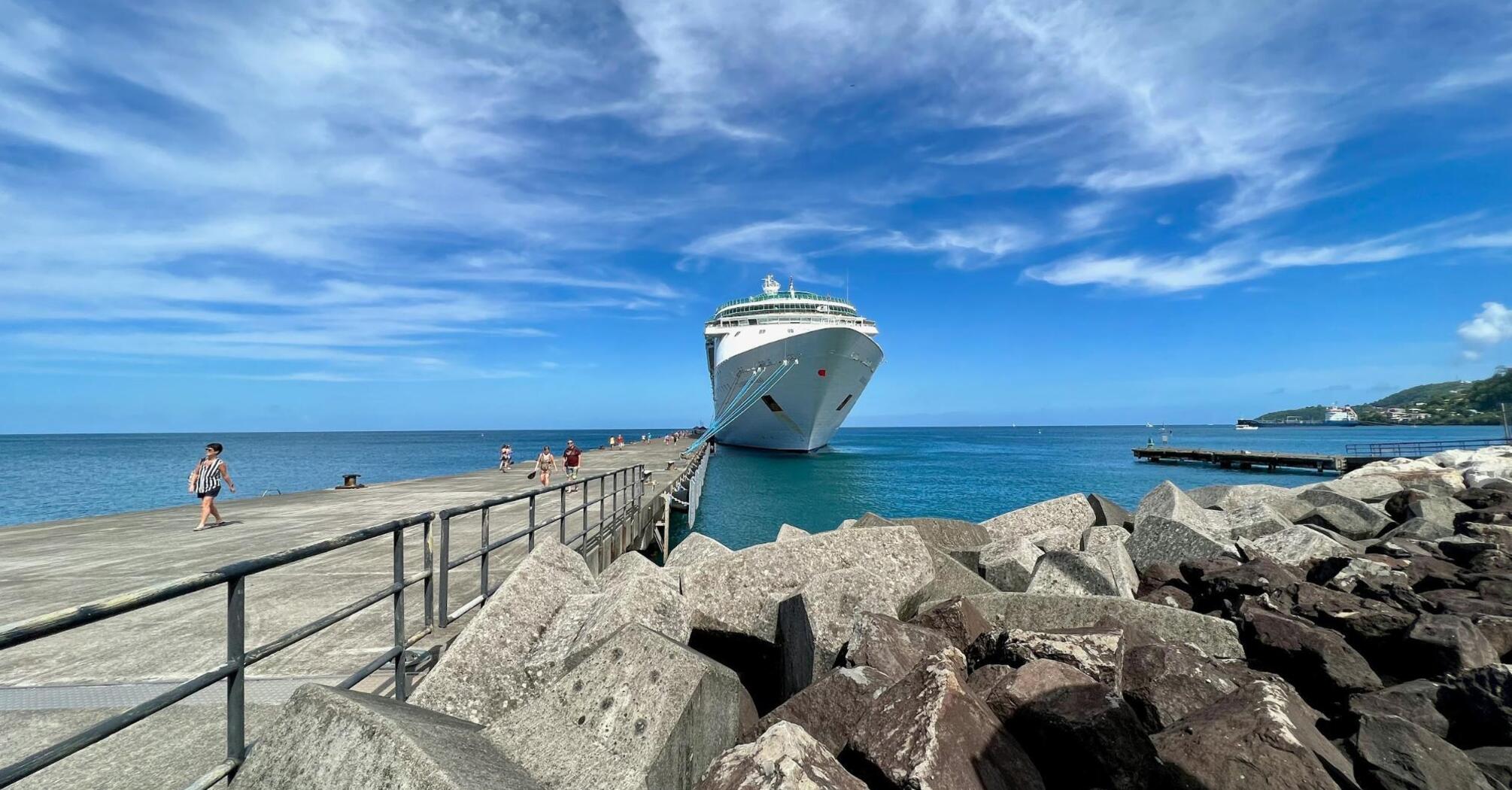 A large cruise ship docked at a pier with people walking along the pier and clear skies overhead