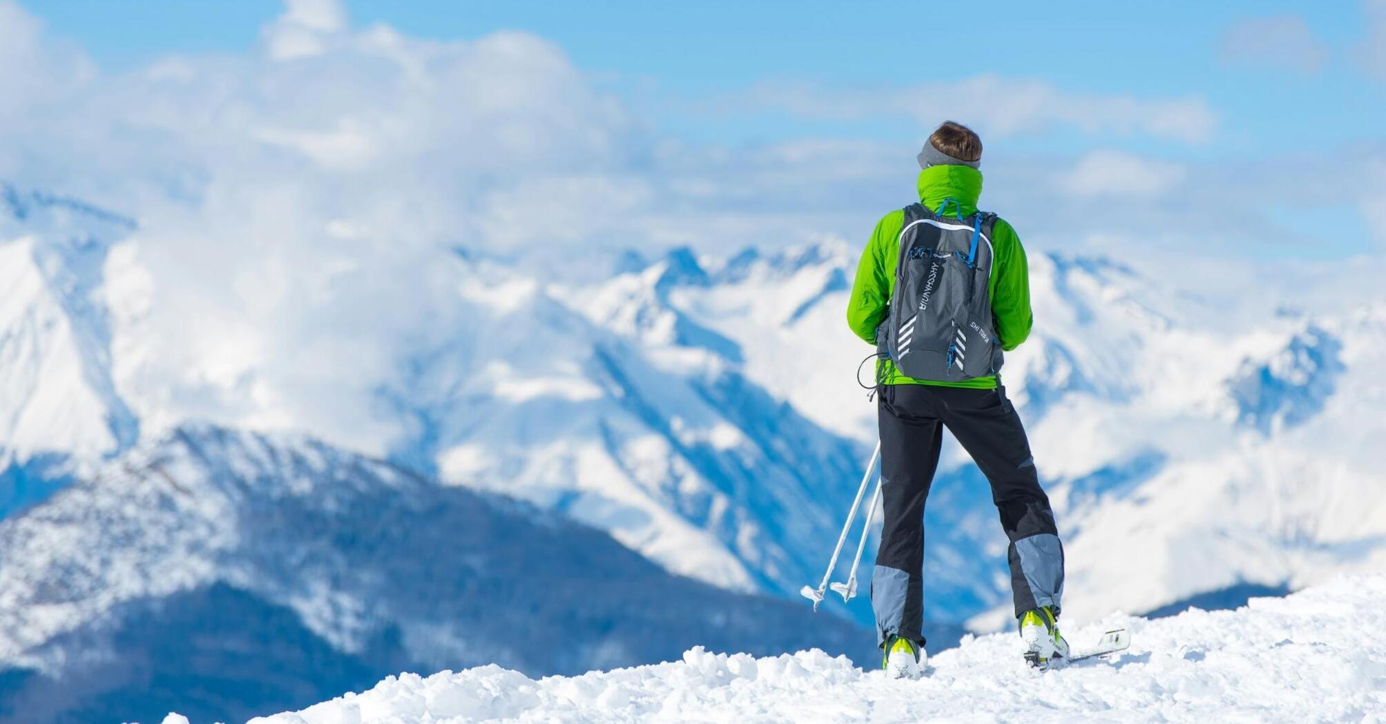 Skier looking at snowy mountain peaks on a sunny day, dressed in green jacket, holding ski poles