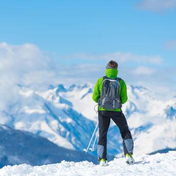 Skier looking at snowy mountain peaks on a sunny day, dressed in green jacket, holding ski poles