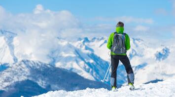 Skier looking at snowy mountain peaks on a sunny day, dressed in green jacket, holding ski poles