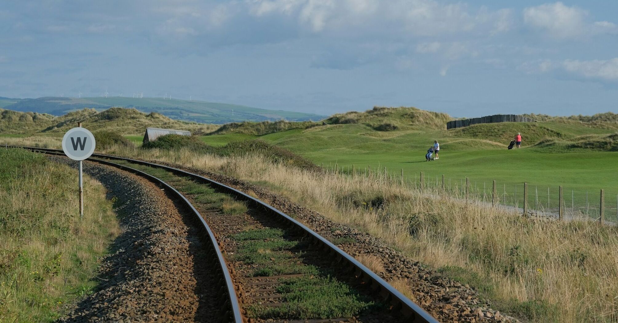 A single-track railway line curves through a grassy landscape, with golfers playing in the distance under a clear sky