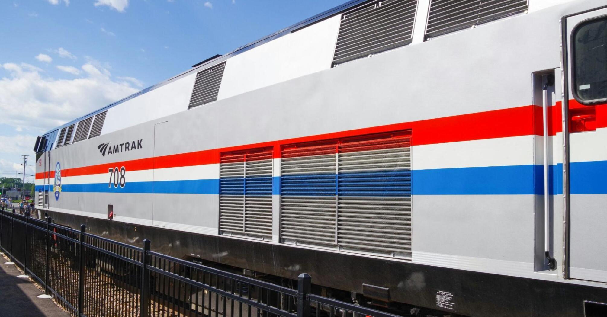 A modern Amtrak train with a red, white, and blue stripe design parked at a station under a clear sky