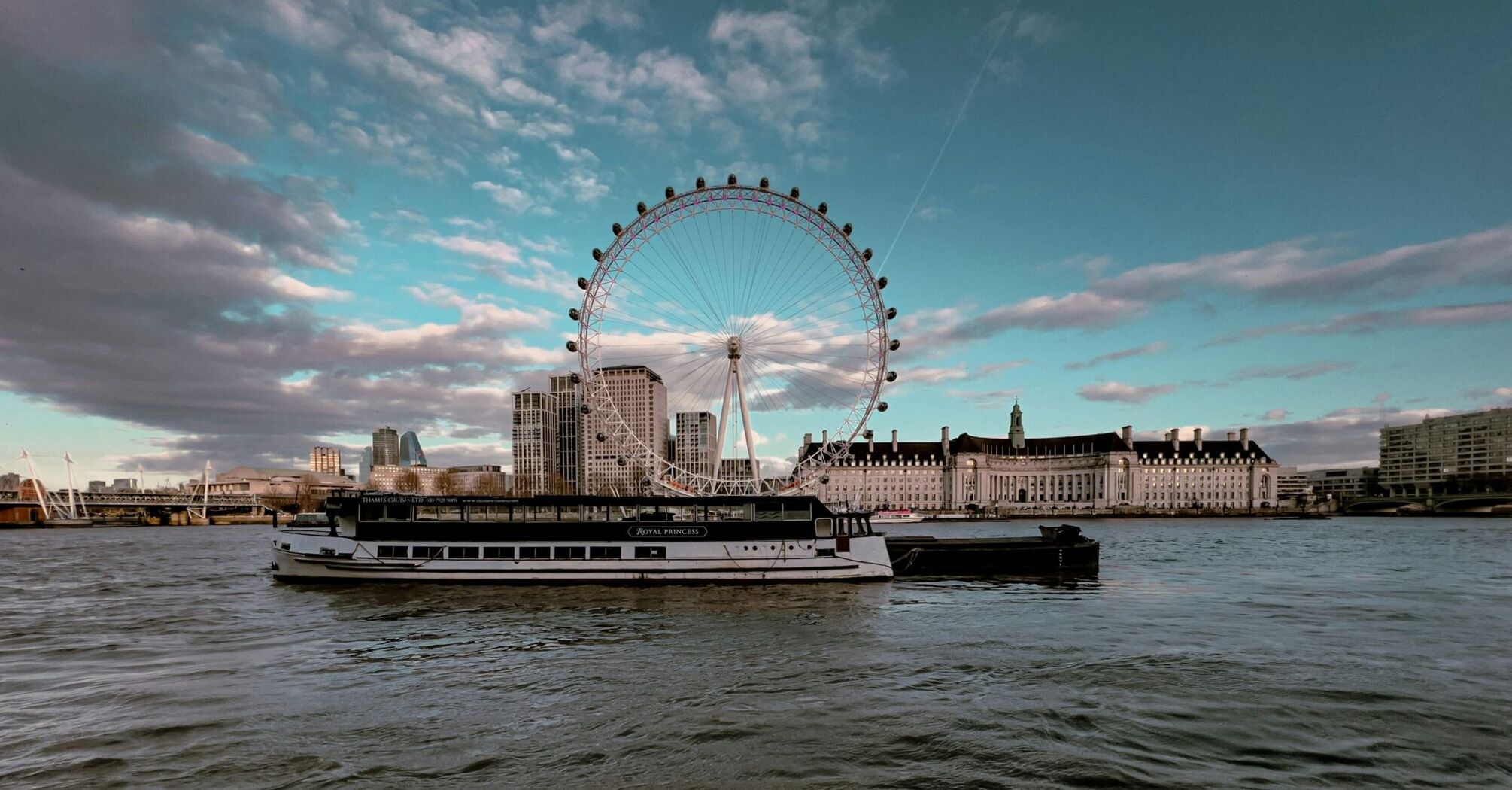 The London Eye on the Thames River with a boat in the foreground under a partly cloudy sky