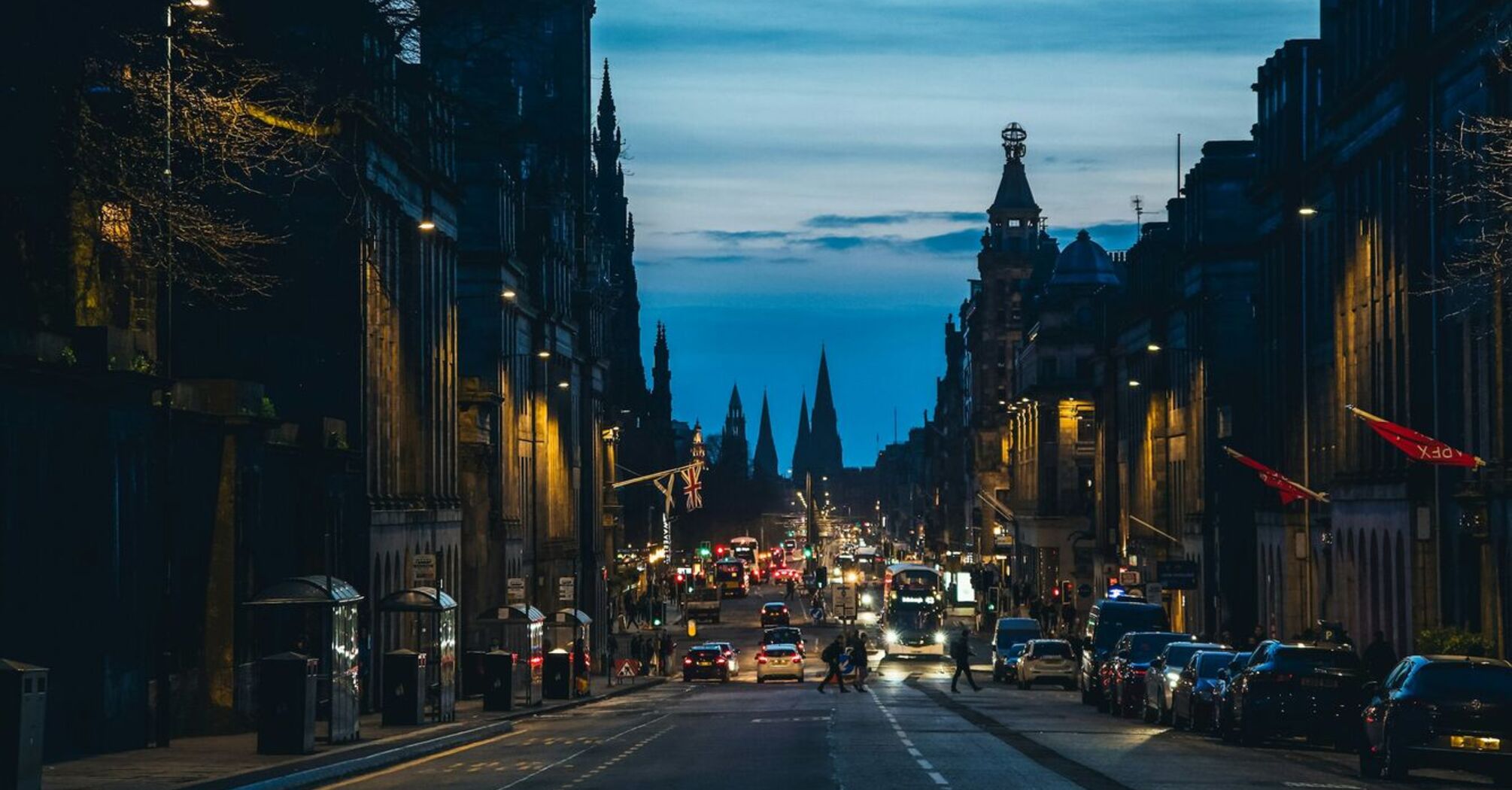 A view of a busy Edinburgh street at dusk, featuring a clock tower illuminated in purple