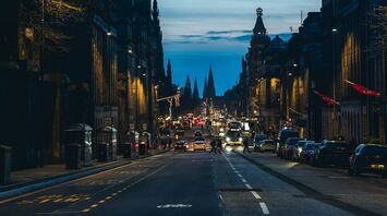 A view of a busy Edinburgh street at dusk, featuring a clock tower illuminated in purple