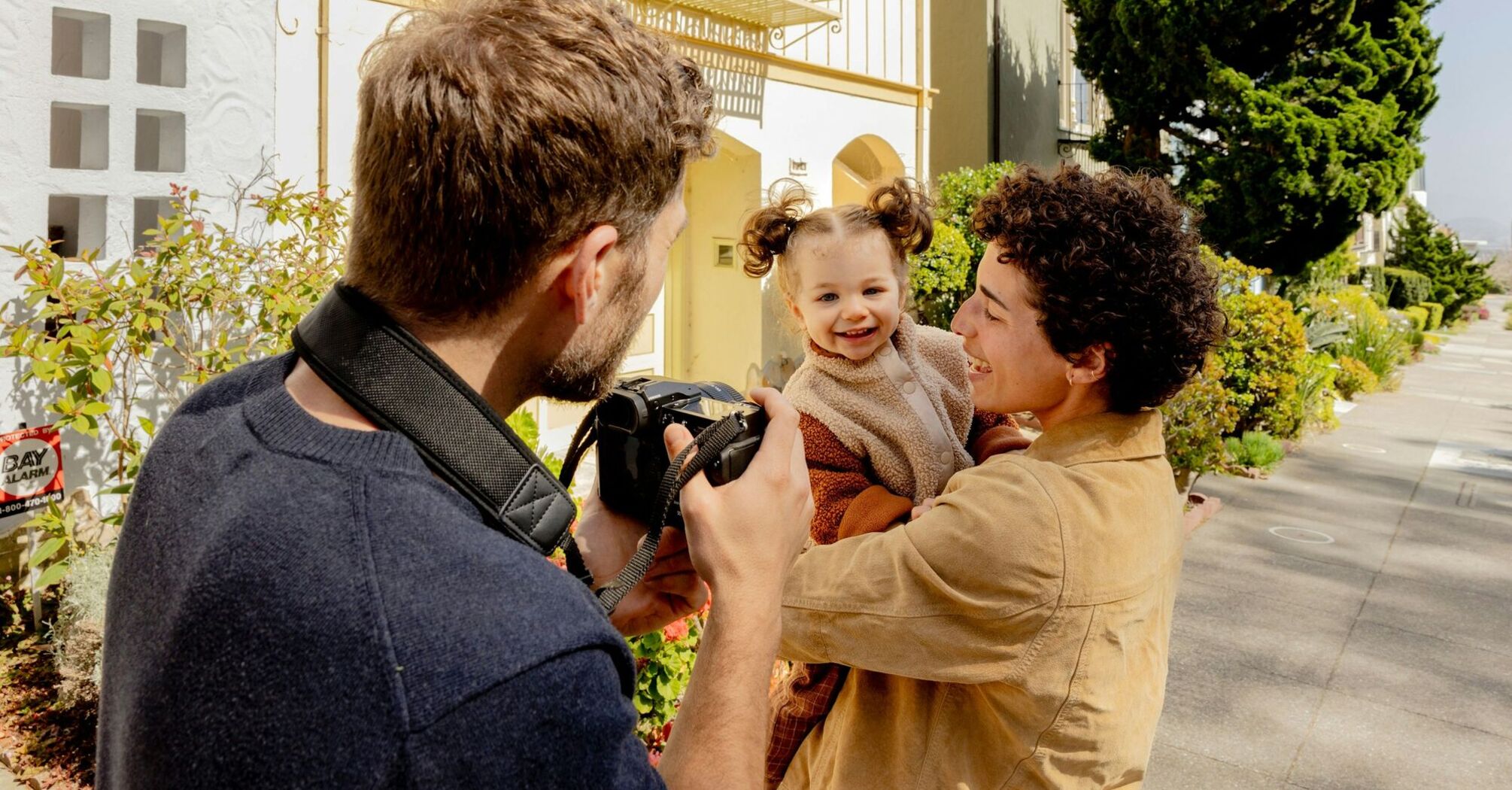 A photographer takes a picture of a smiling child held by an adult outdoors on a sunny day