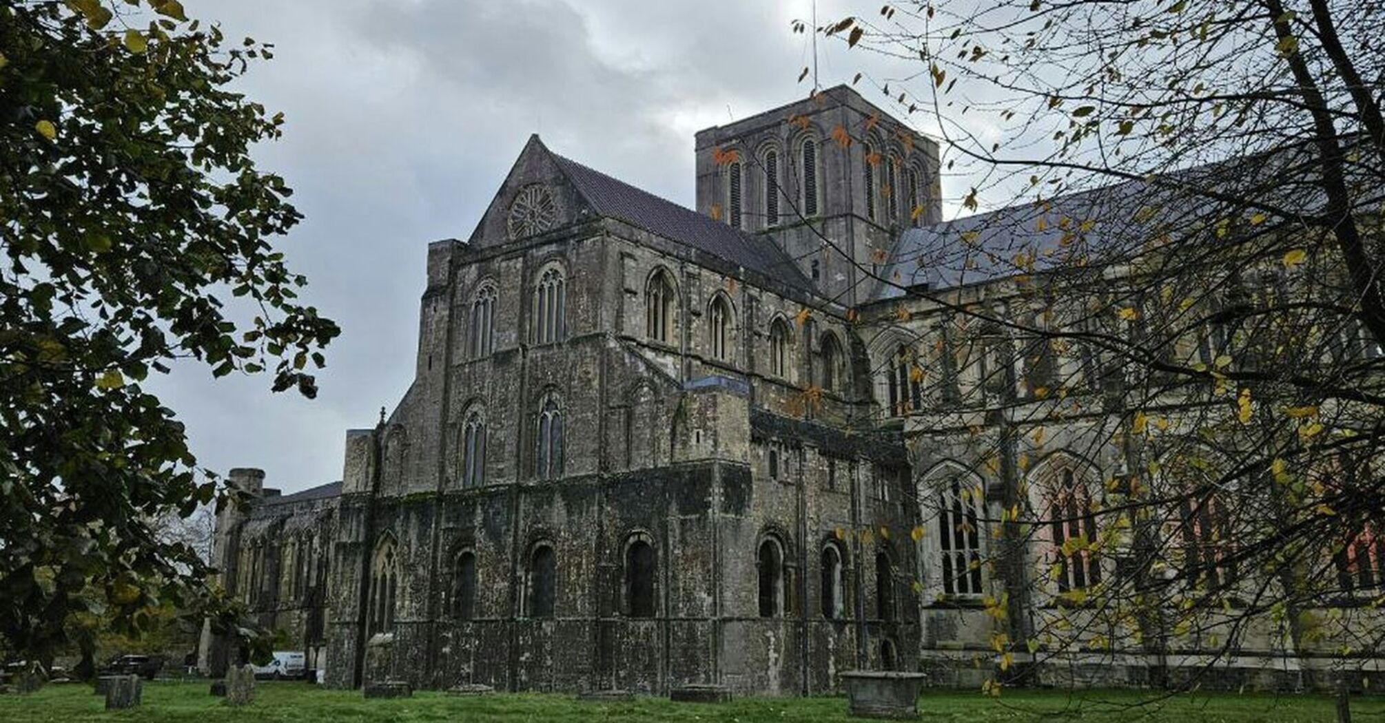 A view of Winchester Cathedral with autumn leaves on the ground and a cloudy sky overhead, framed by tree branches