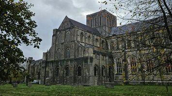 A view of Winchester Cathedral with autumn leaves on the ground and a cloudy sky overhead, framed by tree branches