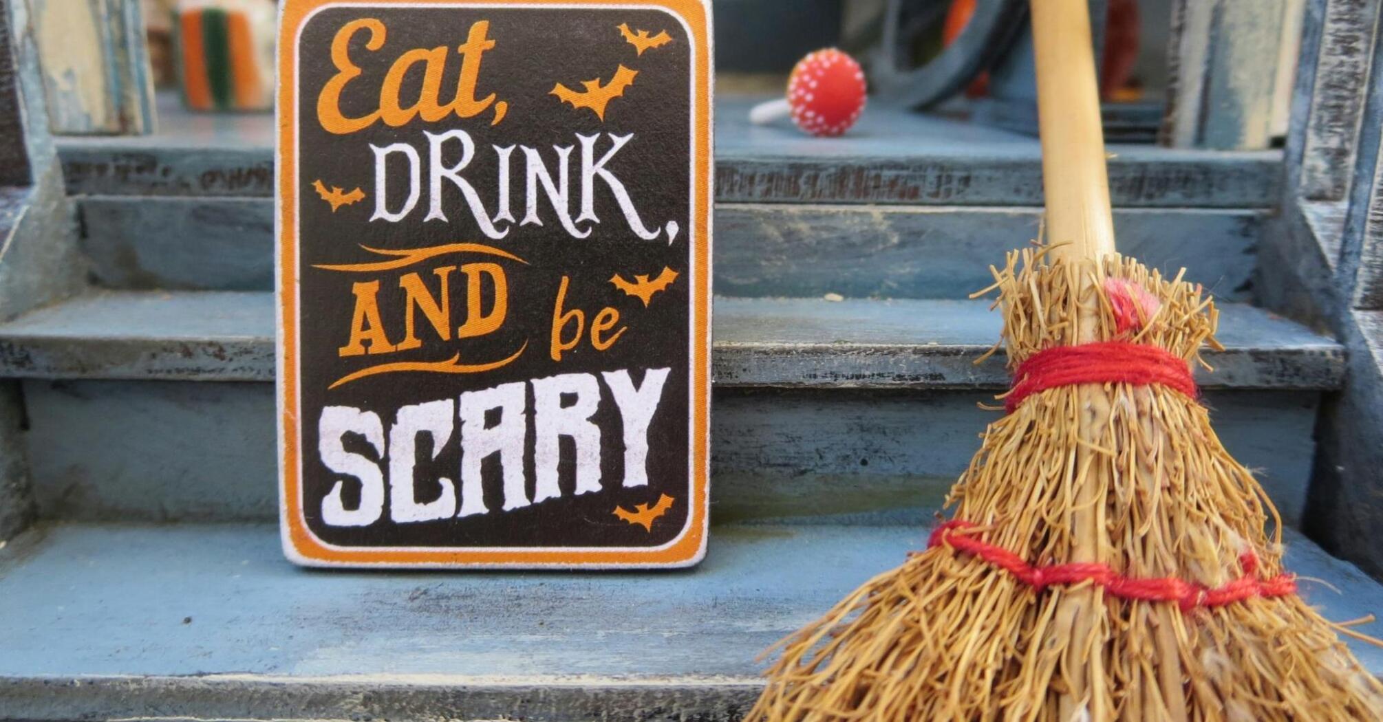 A Halloween-themed sign reading "Eat, Drink, and Be Scary" placed beside a straw broom on steps 