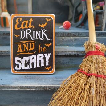A Halloween-themed sign reading "Eat, Drink, and Be Scary" placed beside a straw broom on steps