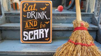 A Halloween-themed sign reading "Eat, Drink, and Be Scary" placed beside a straw broom on steps