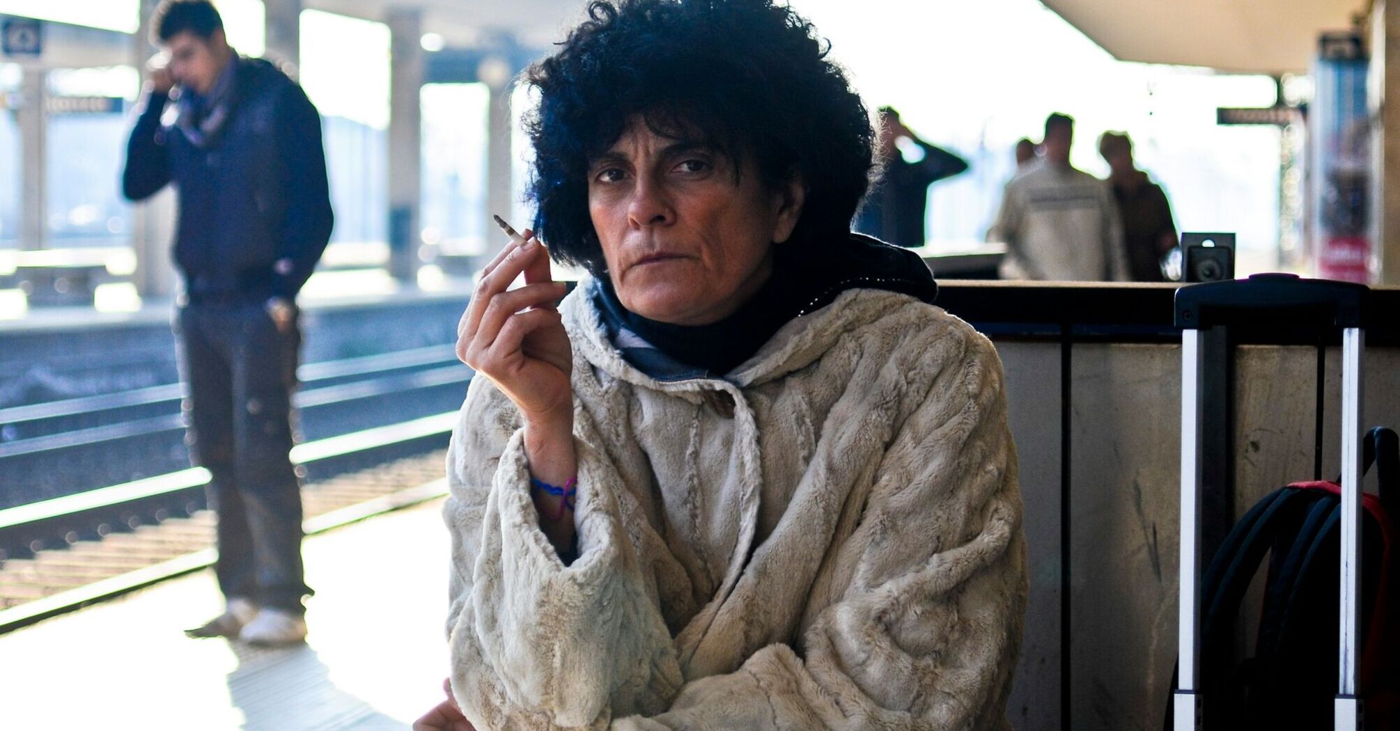 A woman in a heavy coat holding a cigarette, sitting at a train station platform, with a suitcase beside her, waiting while appearing frustrated or thoughtful