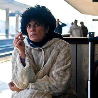 A woman in a heavy coat holding a cigarette, sitting at a train station platform, with a suitcase beside her, waiting while appearing frustrated or thoughtful
