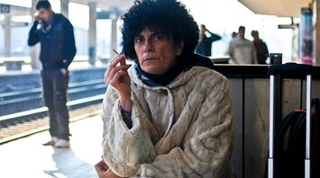 A woman in a heavy coat holding a cigarette, sitting at a train station platform, with a suitcase beside her, waiting while appearing frustrated or thoughtful
