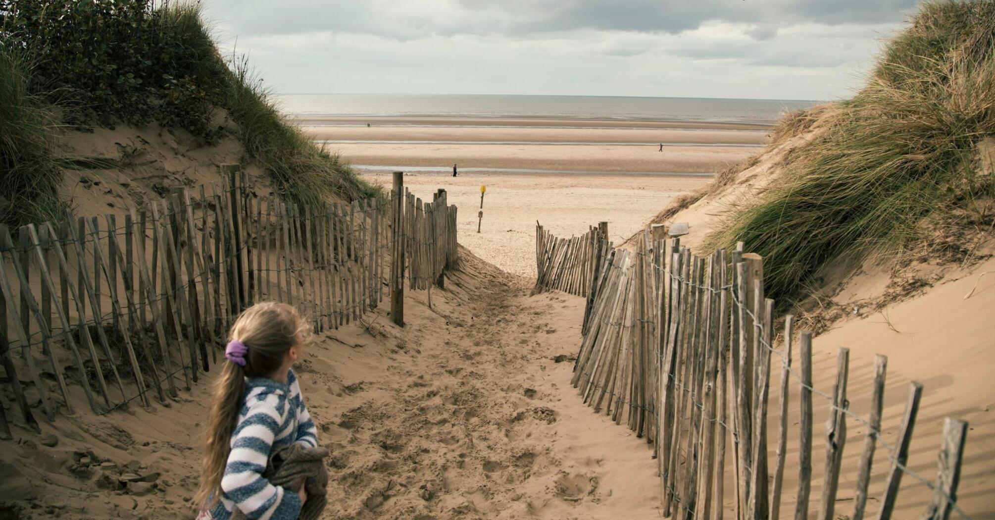 A girl standing on a sandy path leading to a wide, empty beach with sand dunes on both sides under a cloudy sky