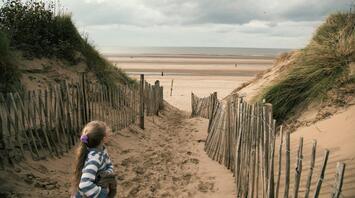 A girl standing on a sandy path leading to a wide, empty beach with sand dunes on both sides under a cloudy sky
