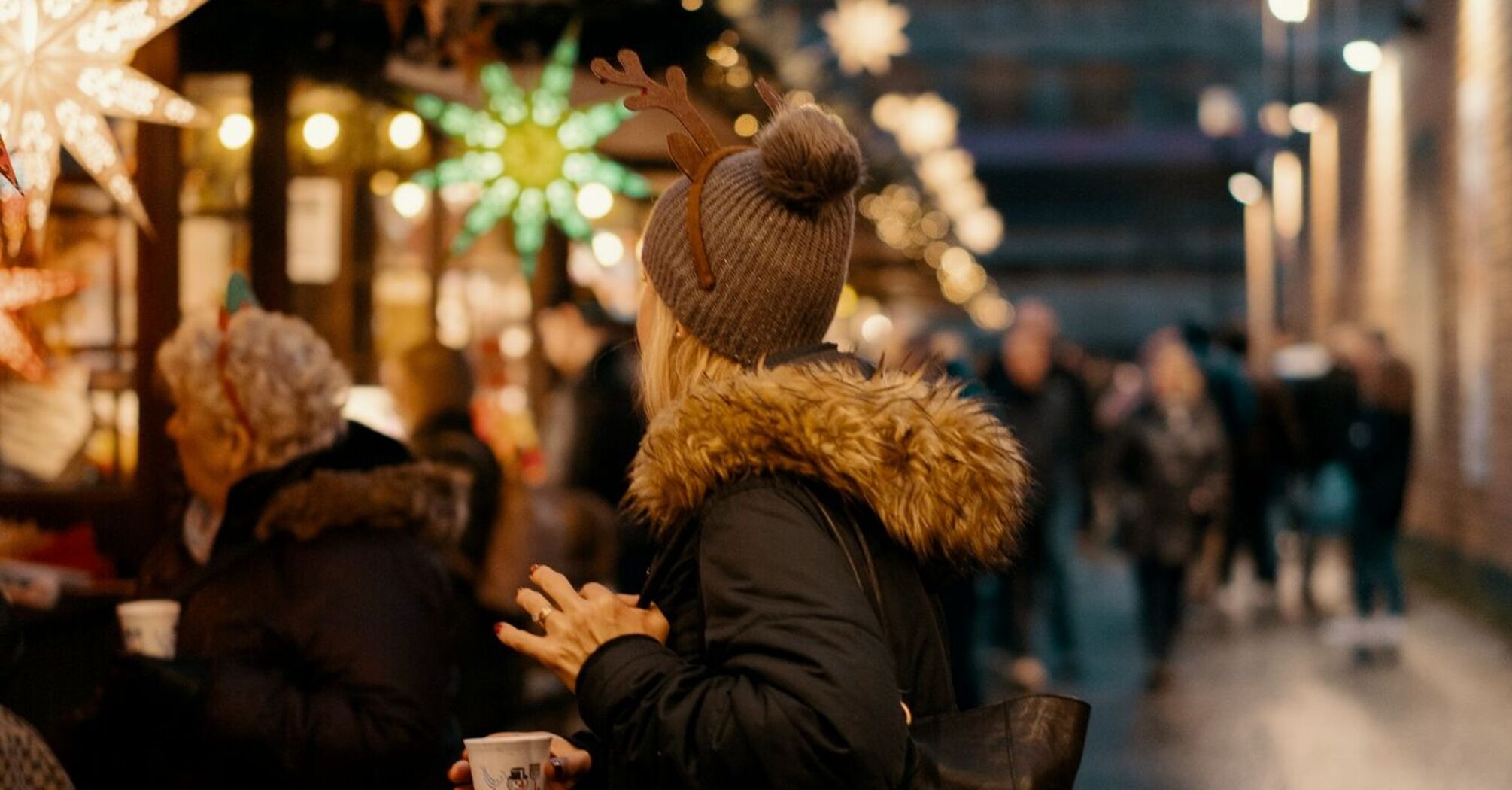 A person in a warm jacket and reindeer antlers stands at a Christmas market stall, holding a cup, surrounded by festive lights and decorations