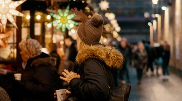 A person in a warm jacket and reindeer antlers stands at a Christmas market stall, holding a cup, surrounded by festive lights and decorations