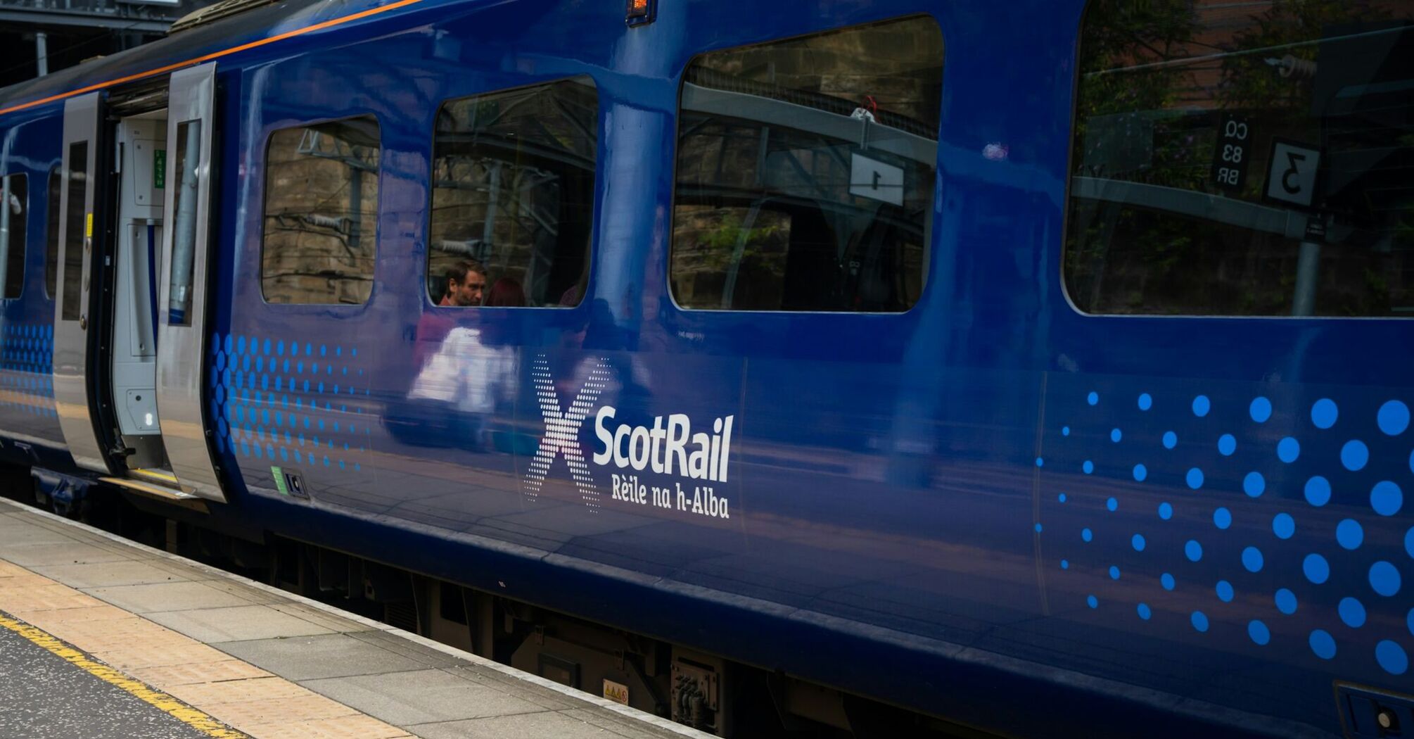 A blue ScotRail train at a platform with passengers visible inside through the window