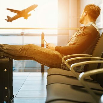 A traveler sitting in an airport lounge with his legs resting on a suitcase, looking out at an airplane taking off