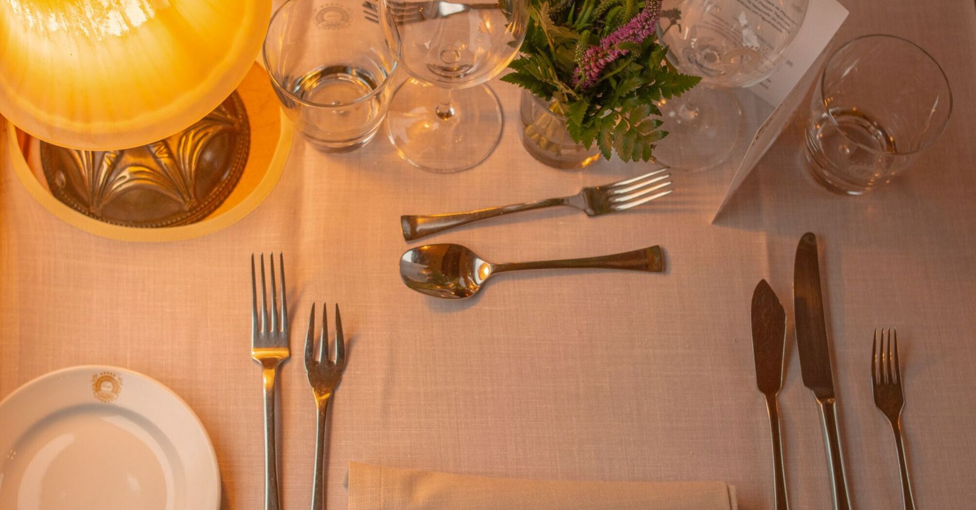 A neatly set table in train with cutlery, glassware, and a small floral arrangement, illuminated by soft lamp lighting