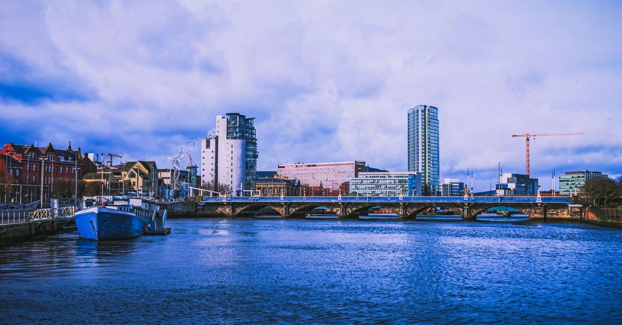 A waterfront view of Belfast City, showcasing modern buildings, a bridge, and a docked boat under a cloudy sky