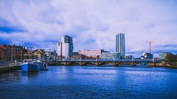 A waterfront view of Belfast City, showcasing modern buildings, a bridge, and a docked boat under a cloudy sky