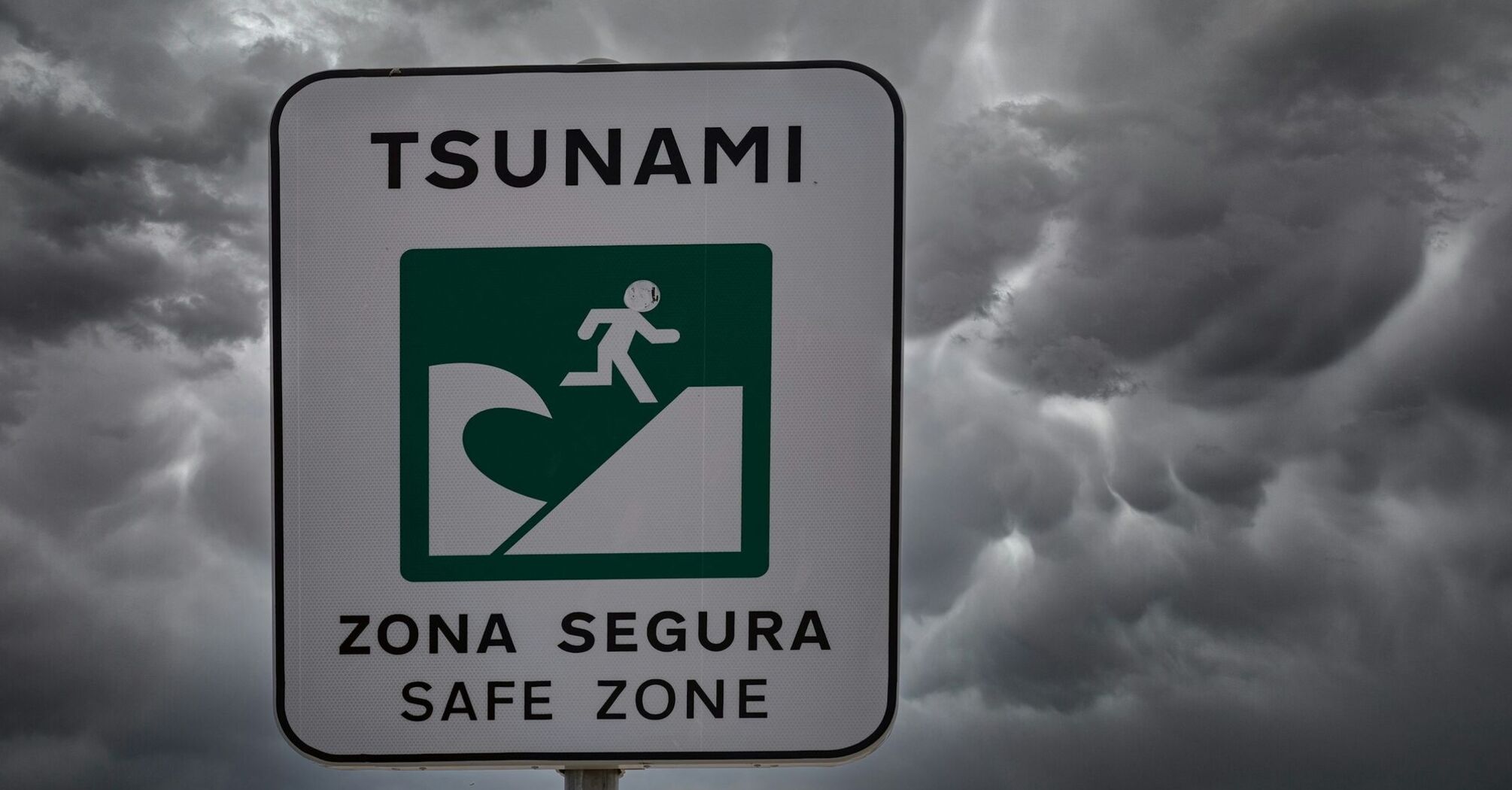 A tsunami evacuation sign reading "Zona Segura - Safe Zone" under a stormy sky, warning of safe areas in case of a tsunami