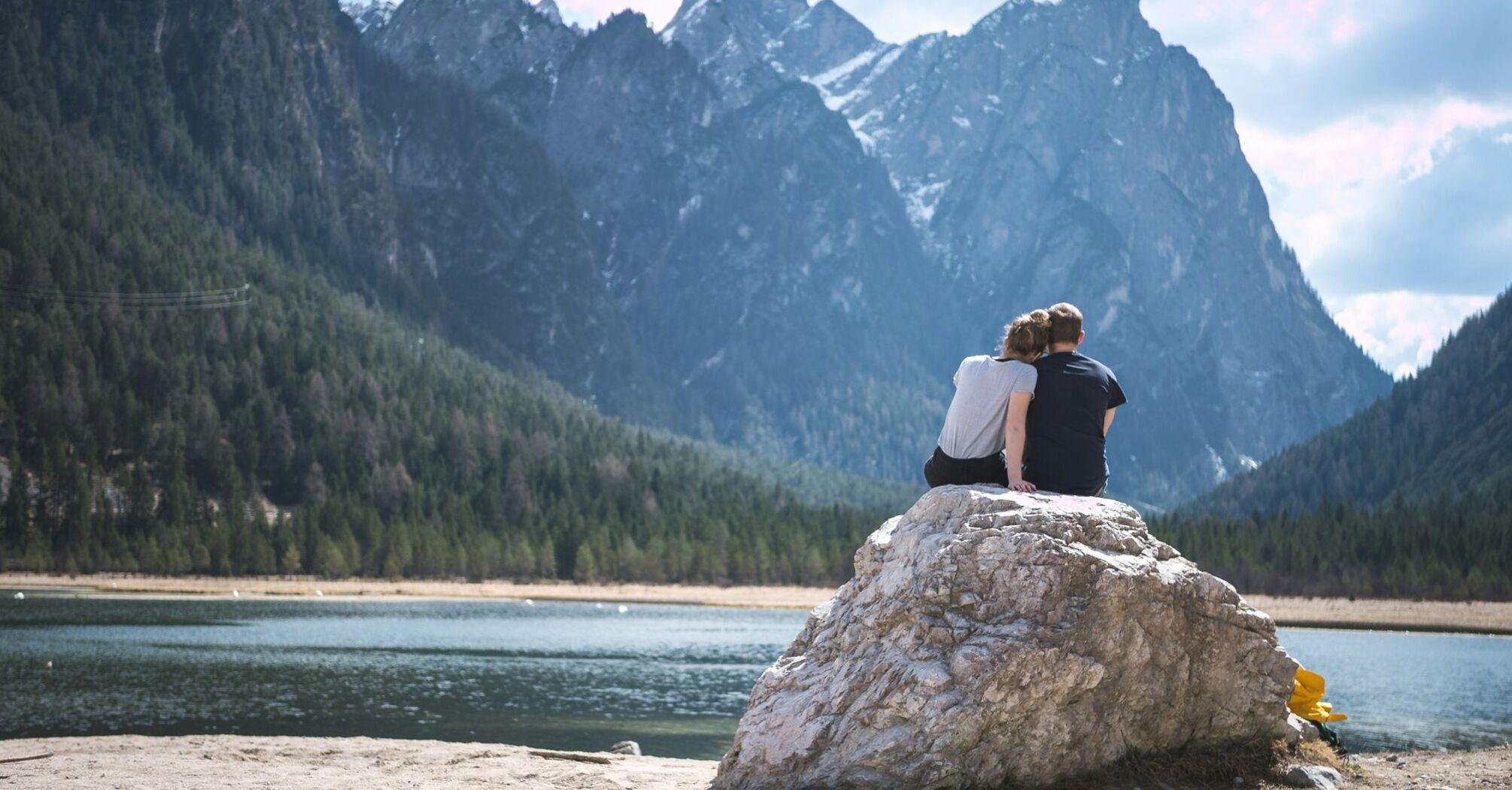 A couple sits on a large rock by a serene mountain lake, surrounded by towering pine trees and high peaks, enjoying a peaceful moment together in nature