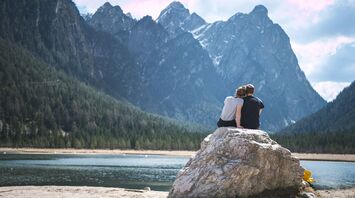 A couple sits on a large rock by a serene mountain lake, surrounded by towering pine trees and high peaks, enjoying a peaceful moment together in nature
