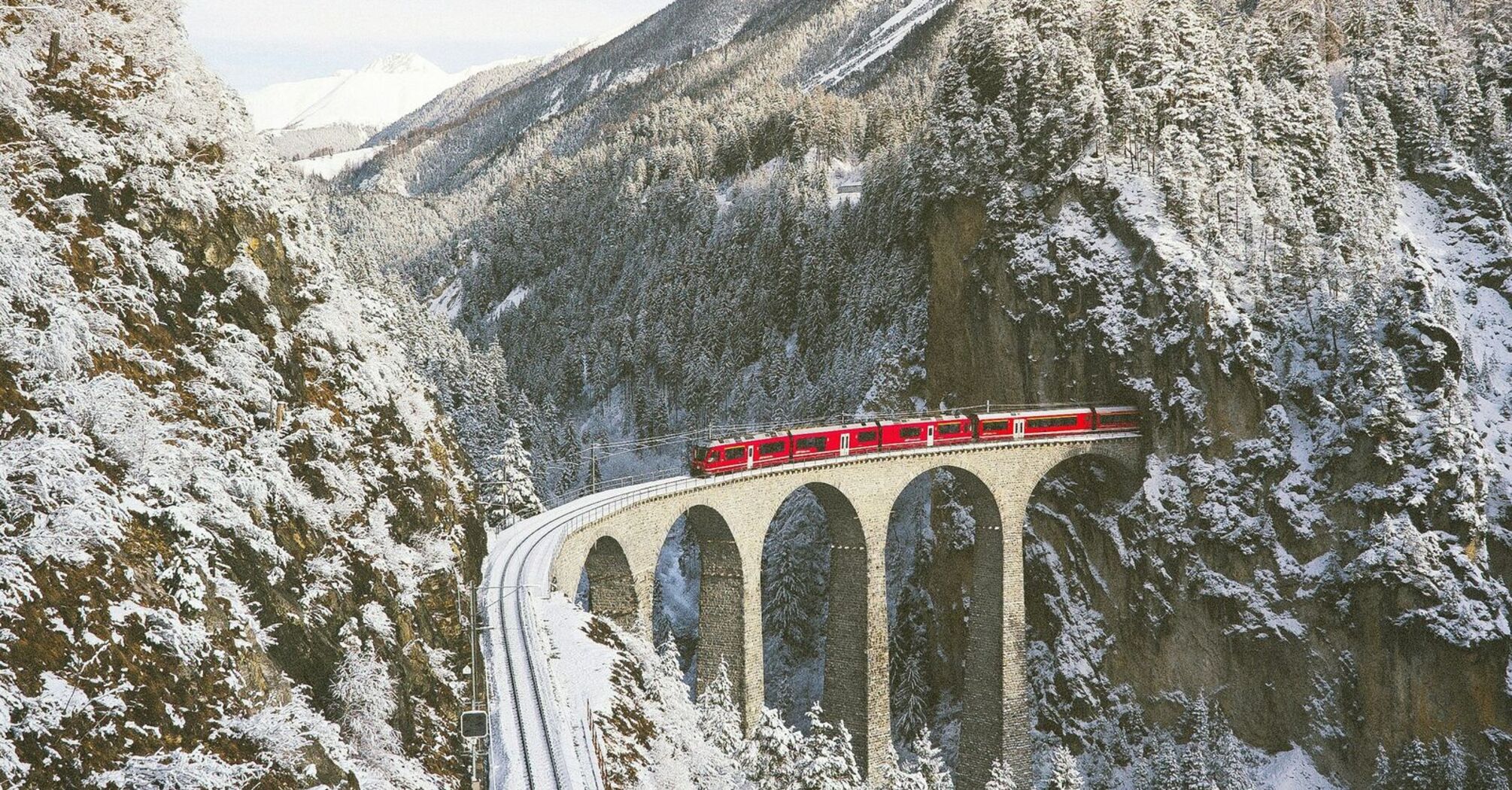 A red train crosses a snow-covered mountain bridge surrounded by winter landscapes