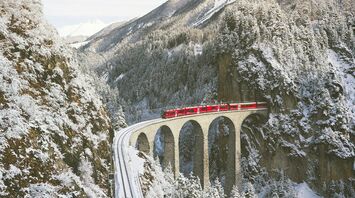 A red train crosses a snow-covered mountain bridge surrounded by winter landscapes