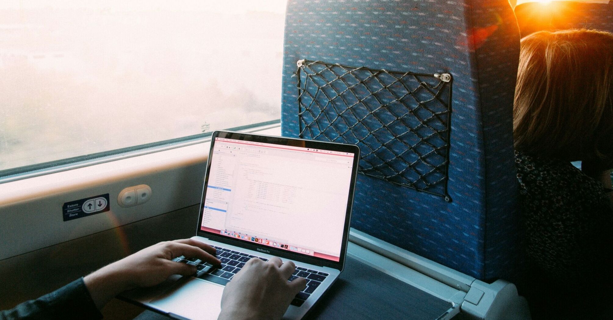 A person working on a laptop while traveling by train, with a view of the outside through the window as the sun sets