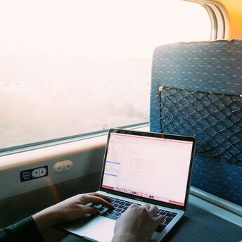 A person working on a laptop while traveling by train, with a view of the outside through the window as the sun sets
