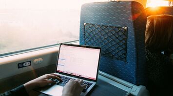 A person working on a laptop while traveling by train, with a view of the outside through the window as the sun sets