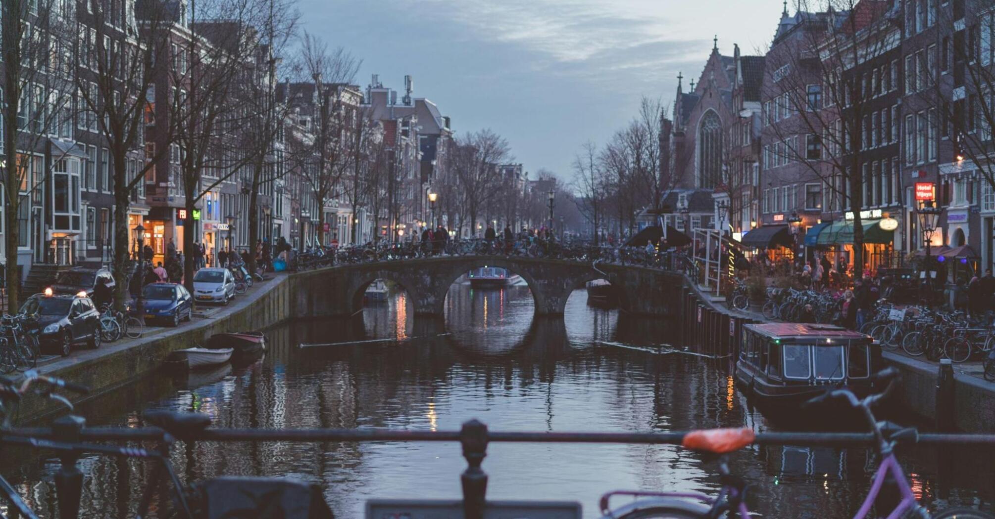 Evening view of an Amsterdam canal with a bridge and city lights reflecting in the water, framed by bicycles in the foreground